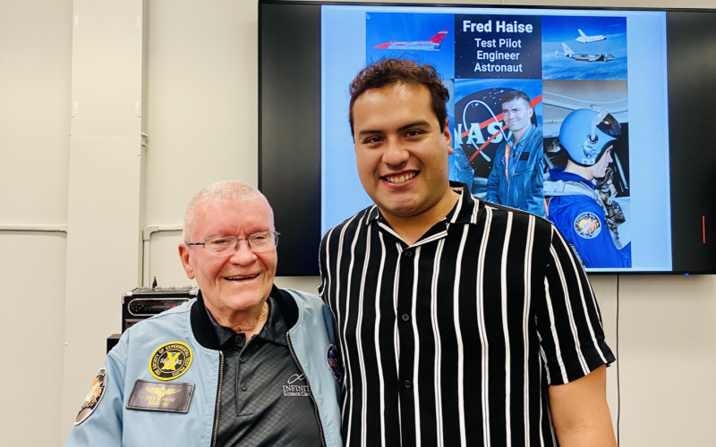 A young man in a pinstripe shirt stands next to an older man in a blue space jacket as they smile for a photo. Behind them a screen reads "Fred Haise test pilot engineer astronaut"