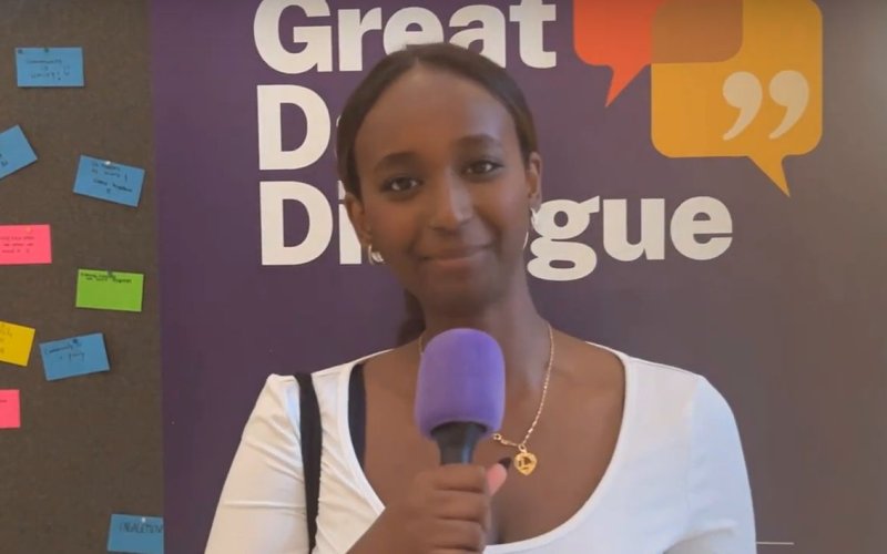 a young woman holding a microphone stands in front to a sign that reads Great Dane Dialogue