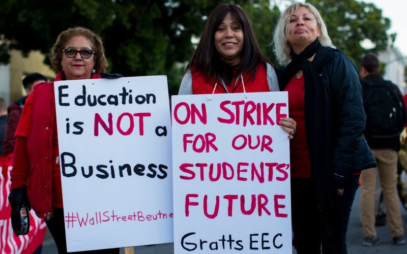 Three women, two of them holding signs that includ the words "Education is NOT a Business" and "On strike for our students' future"