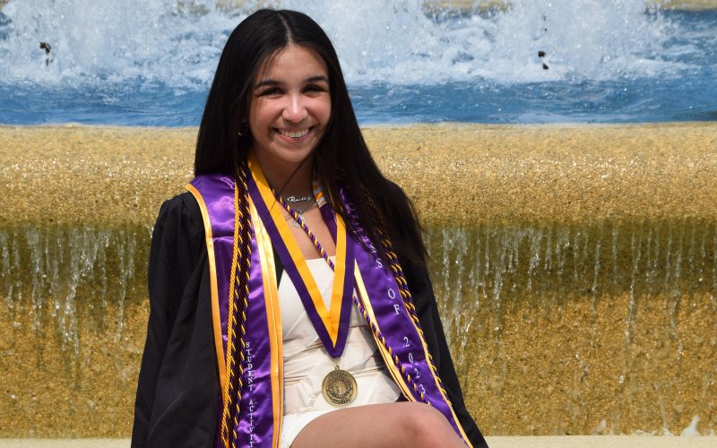 UAlbany graduate Reilly Coleman, dressed in her graduation robes, sits in front of a fountain at UAlbany.