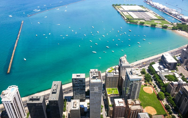 Aerial image of the Chicago shoreline along Lake Michigan. The bottom third of the image shows city buildings with tree lined streets and two baseball diamonds. The top two-thirds of the image is aqua blue water, dotted with boats, two jetties and two fingers of land reaching out into the lake. One has an amusement park and Ferris wheel. The other has a mix of parkland and water purification infrastructure. 