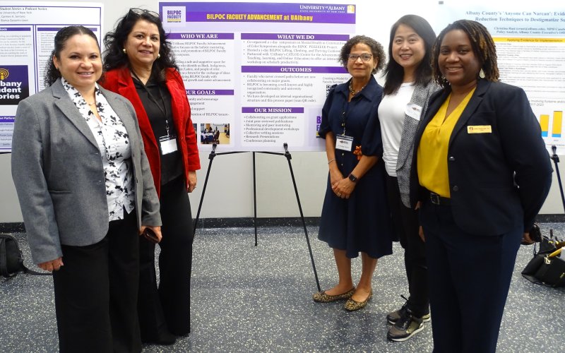 Five women in professional attire stand, smiling, in front of a purple poster set on a black tripod. The poster is titled 'BILPOC Faculty Advancement Initiative at UAlbany' and has subheads reading: who we are, our goals, what we do, outcomes, and our mission.