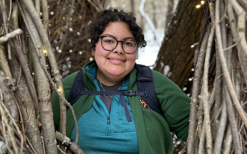 A young women with dark curly hair and glasses wearing a green fleece coat and backpack stands, smiling, inside a rustic structure made of interwoven twigs and fairy lights with snow in the foreground. 