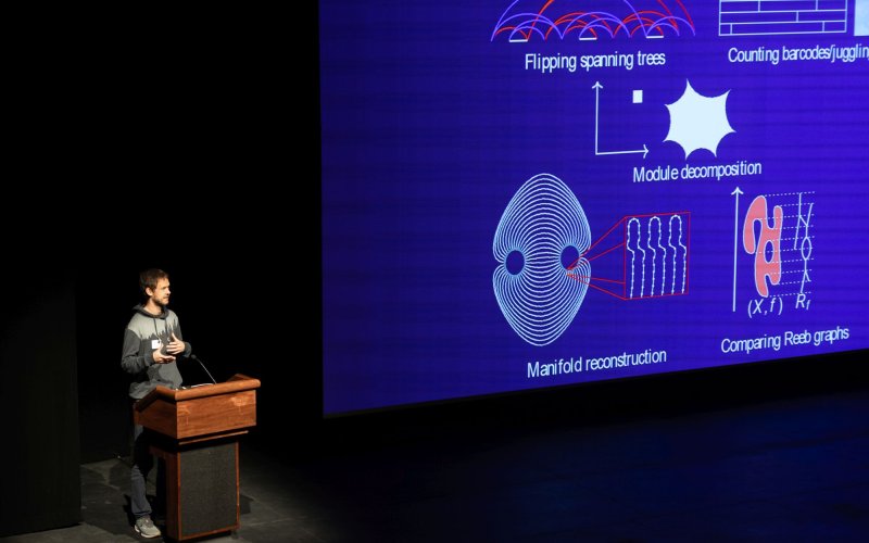 A man speaks from a podium next to a projection of mathmatical images.