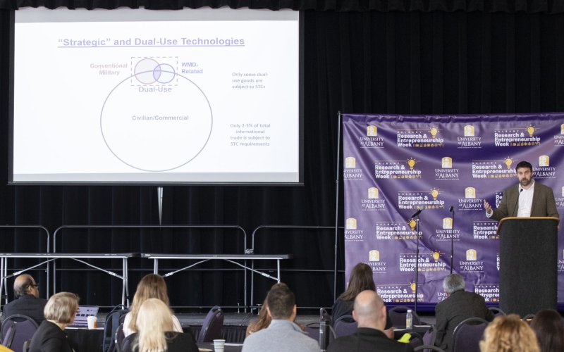 A man speaks to an audience from a podium, standing in front of a purple cloth reading "research and entrepreneurship week" and next to a screen with the words "strategic and dual-use technologies" 