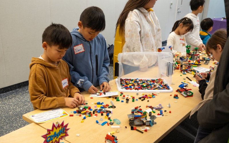 Children work on projects at a table scattered with Lego pieces.