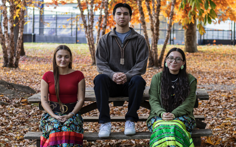 Two young women in long colorful skirts and a young man sit at a picnic table on the UAlbany campus on a sunny fall day. Colorful leaves decorate the trees and ground.