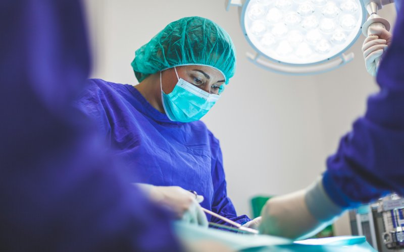 An oral surgeon wearing blue scrubs and teal PPE works on a patient in a brightly lit medical office.