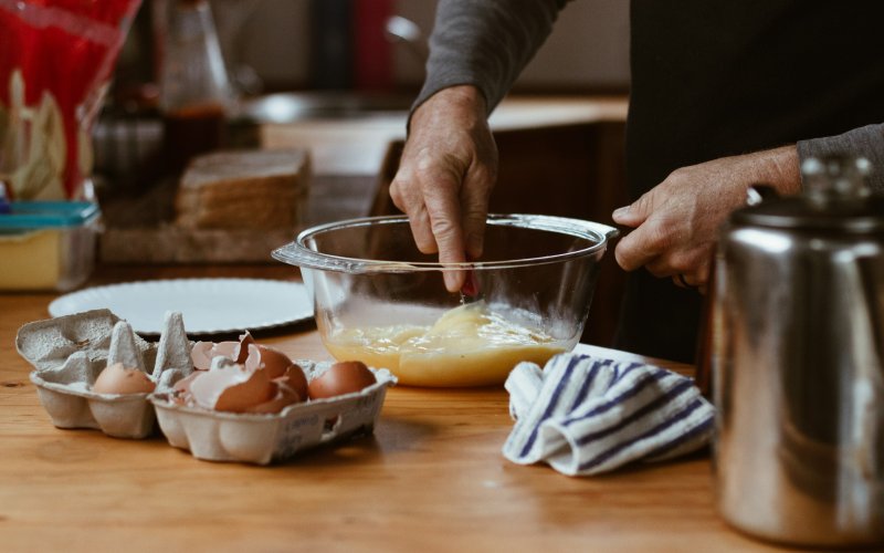 Various materials for cooking are arranged on a wooden kitchen countertop. A half-dozen egg carton is open and full of cracked eggshells. A man whisks the eggs in a glass bowl with bare hands.. 