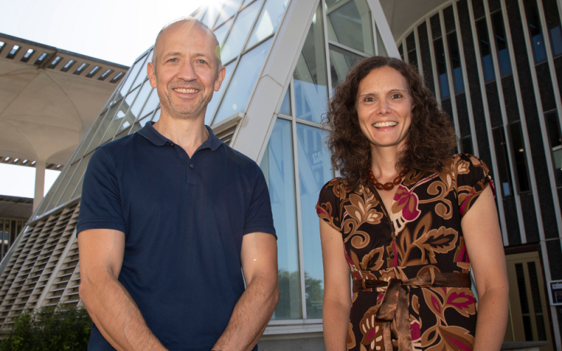 Drs. Yucel and Friedman stand smiling at the camera on the main UAlbany campus with glass architecture in the background.