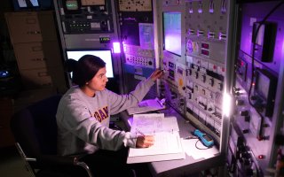 A student sits at a computer terminal with multiple screens and dials in a physics lab.