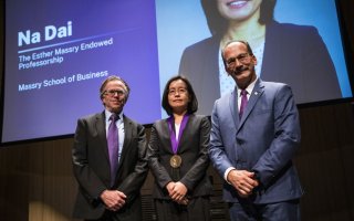 A woman in a gray suit, wearing a medalion, stands between two men in front of a screen with her photo and the words "Na Dai, the Esther Massry Endowed Professorship, Massry School of Business"