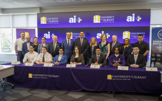 Chancellor King, along with SUNY leaders and elected officials, stand behind a group of eight UAlbany students sitting at a table branded with the UAlbany Minerva logo.