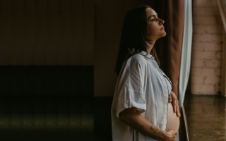 A women in an open nightshirt, hands on her pregnant belly, stares out a window.