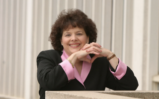 A woman with short curly hair in a pink and black blazer clasps her hands together and rests her elbows on a concrete ledge, smiling for a portrait.