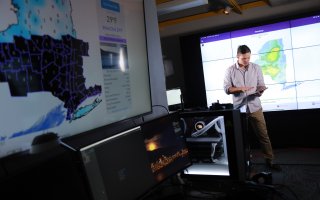 A man with an ipad looks down while a weather map of NEw York state appears in the foreground at UAlbany's XCite Lab.