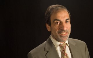 A man with short gray hair wearing a tan suit and gold tie with red stripes smiles for a portrait against a black backdrop.