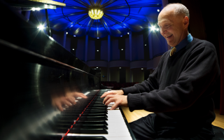 Closeup of a smiling man with short gray hair wearing a black sweater playing piano on a lit-up stage