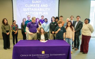 A group of smiling people stand around a woman signing a document while seated at a table with a purple cloth reading "Office of Environmental Sustainability"