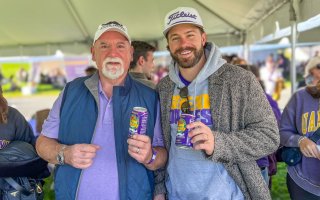 Two men with beards and white ball caps stand in a tend holding purple cans of Reed's ginger beer, decorated with a white mustache.