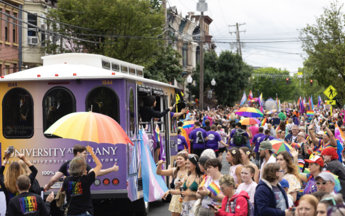 Provost Carol Kim waves to a crowd of onlookers from inside a purple and gold UAlbany trolley at the Capital Pride Parade and Festival in Albany. Throngs of people in colorful outfits and holding rainbow umbrellas fill the street.