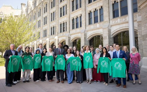 A line of people standing in front of the SUNY Administration building hold green long-sleeved T-shirts with a round white logo