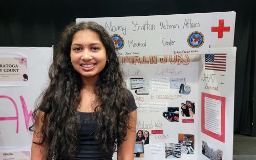 A smiling young woman with long dark curly hair stand in front of her informational poster about her internship at a VA hospital.