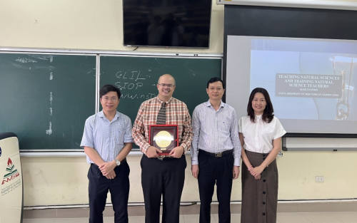 Four people pose for a picture in front of a chalkboard and video screen in a classroom. One of them holds a plaque.