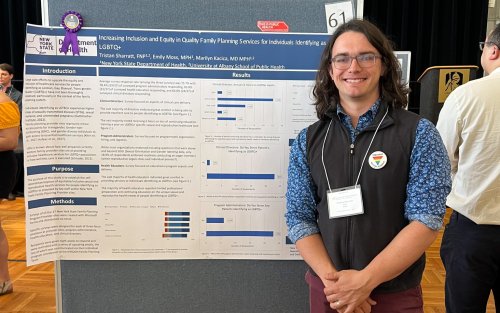 A young man with shoulder length brown hair and glasses smiles next to a blue and white poster pinned to a gray display board in UAlbany's ballroom during the School of Public Health poster symposium in May 2024.