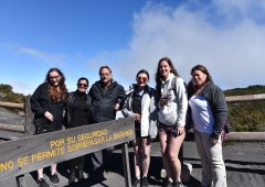 A small group of students and a faculty member stand beside a sign in Spanish. They are outdoors and a clear blue sky is behind them.