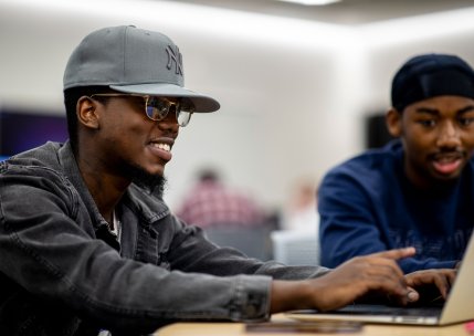 Two students smile as they sit together at a table looking at laptops.
