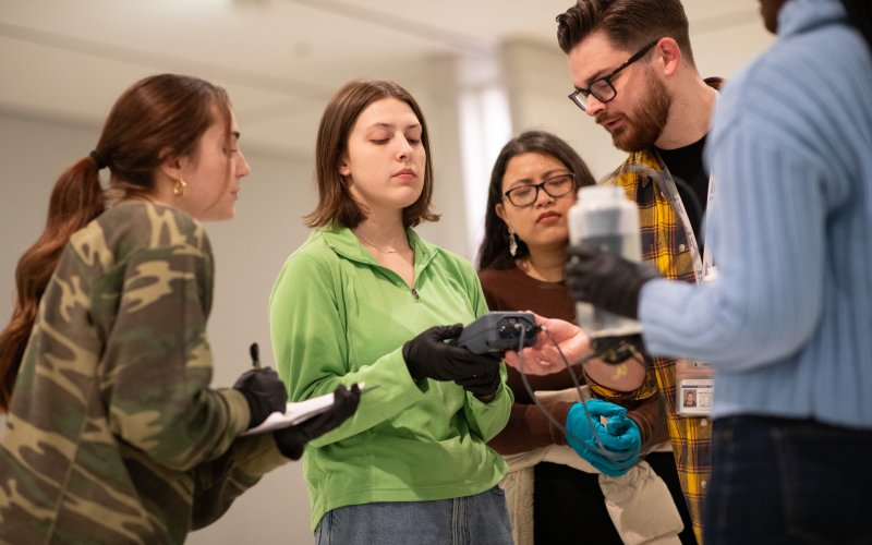 Five young people, four women and one man, stand together holding instruments to collect and record water chemistry measurements. A woman in the center is holding a gray electronic device connected to wires with probes placed in a water bottle held by a woman wearing a blue sweater and black gloves. A woman wearing a camo print shirt holds a clipboard and looks on. A woman in a brown sweater and a man in a yellow plaid shirt also look on. 