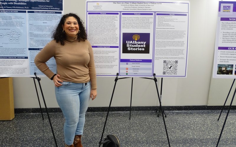 A young woman with curly brown hair wearing a tan sweater and brown boots smiles in front of a poster about the UAlbany Student Stories Podcast.