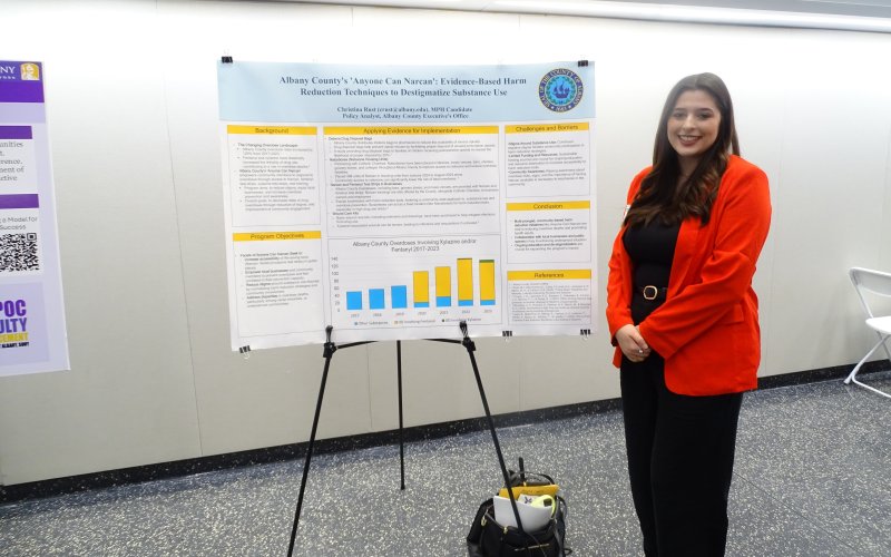 A young woman with long dark hair, wearing a red blazer and black pants smiles beside her poster titled "Albany County's 'Anyone Can Narcan': Evidence based harm reduction techniques to destigmatize substance use." 