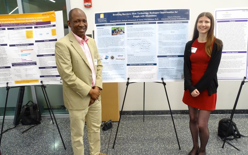 A man in a tan suit and a woman in a red dress with black cardigan smile in front of their poster titled "Breaking Barriers: How Technology Reshapes Opportunities for People with Disabilities."