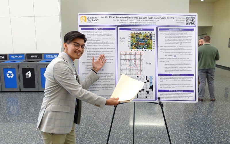 A young man wearing glasses and a grey blazer displays his poster titled "Healthy Minds and Emotions: Evidence Brought Forth from Puzzle Solving."