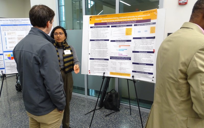 A woman in a striped scarf and glasses stands in front of a poster, speaking to a man in a grey jacket. Her poster reads: “Epigenetic mechanisms linking childhood adversity to substance use disorder in young adulthood from the ALSPAC Study."