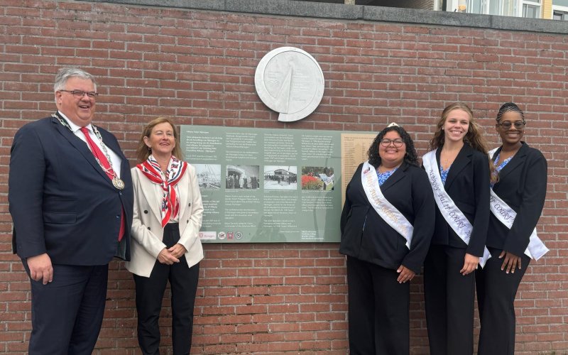 Five people, one man and four women, wearing business attire stand smiling in front of a brick wall bearing an informational panel and large metal seal. 