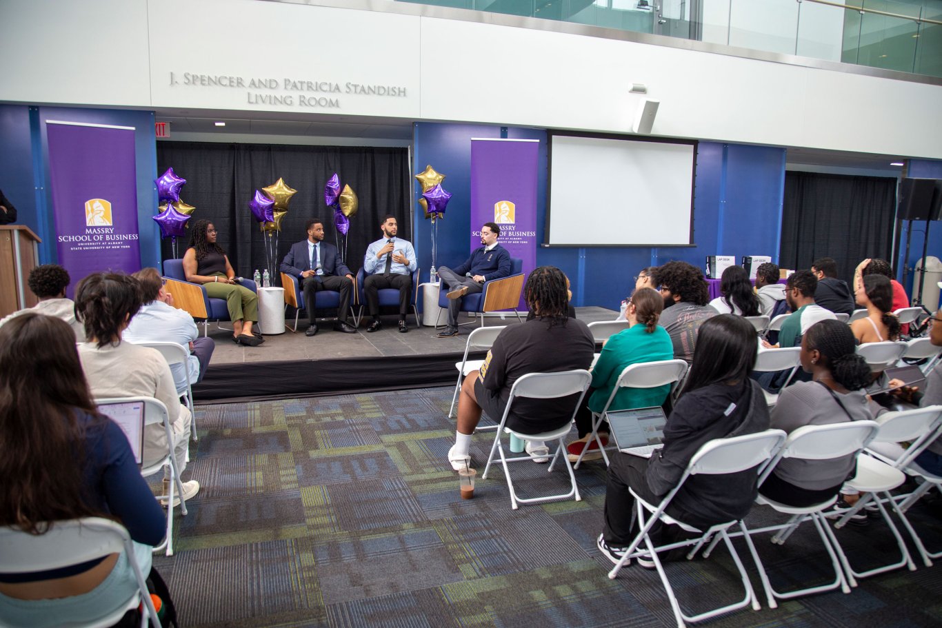 Four students sit on a stage, with one speaking into a microphone as a seated audience listens on Showcase Day.
