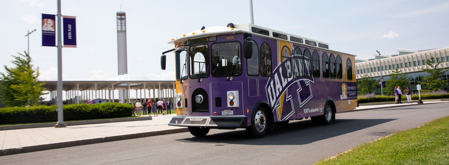An electric trolley decorated with UAlbany logos and colors parked on Collins Circle on the Uptown Campus, with the carillon in the background.