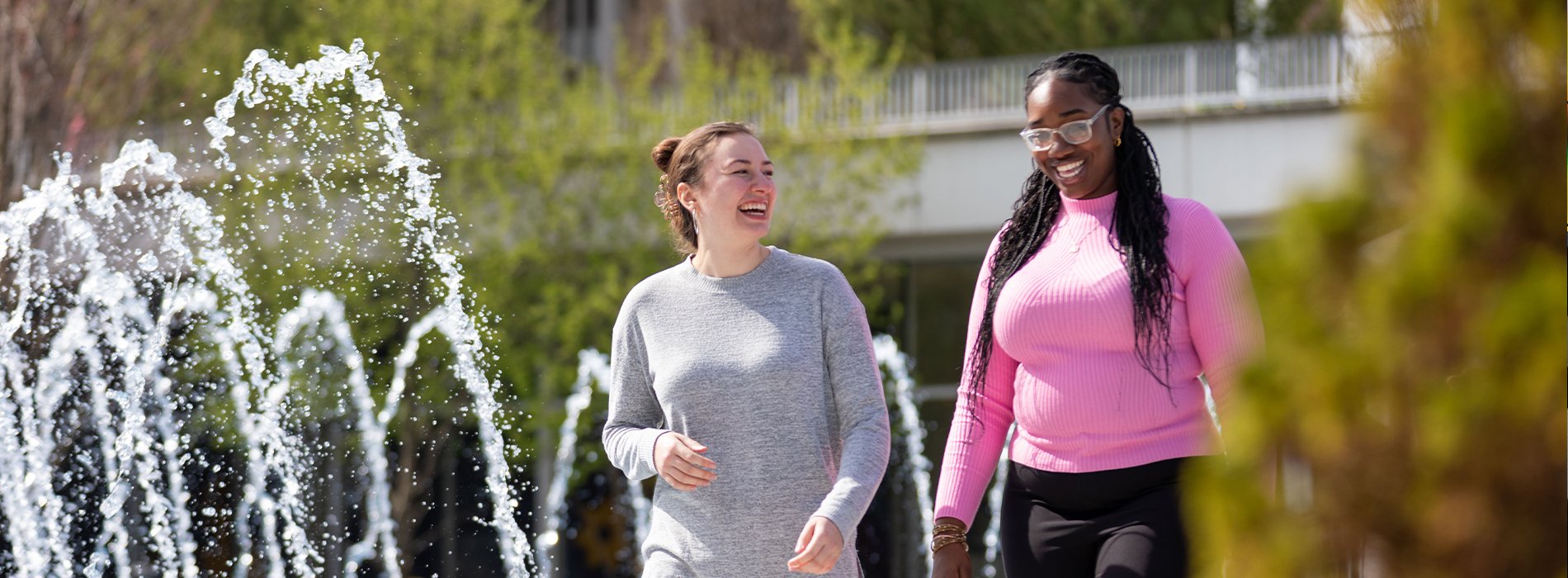 Two UAlbany students smile as they walk by a fountain on campus on a sunny day.