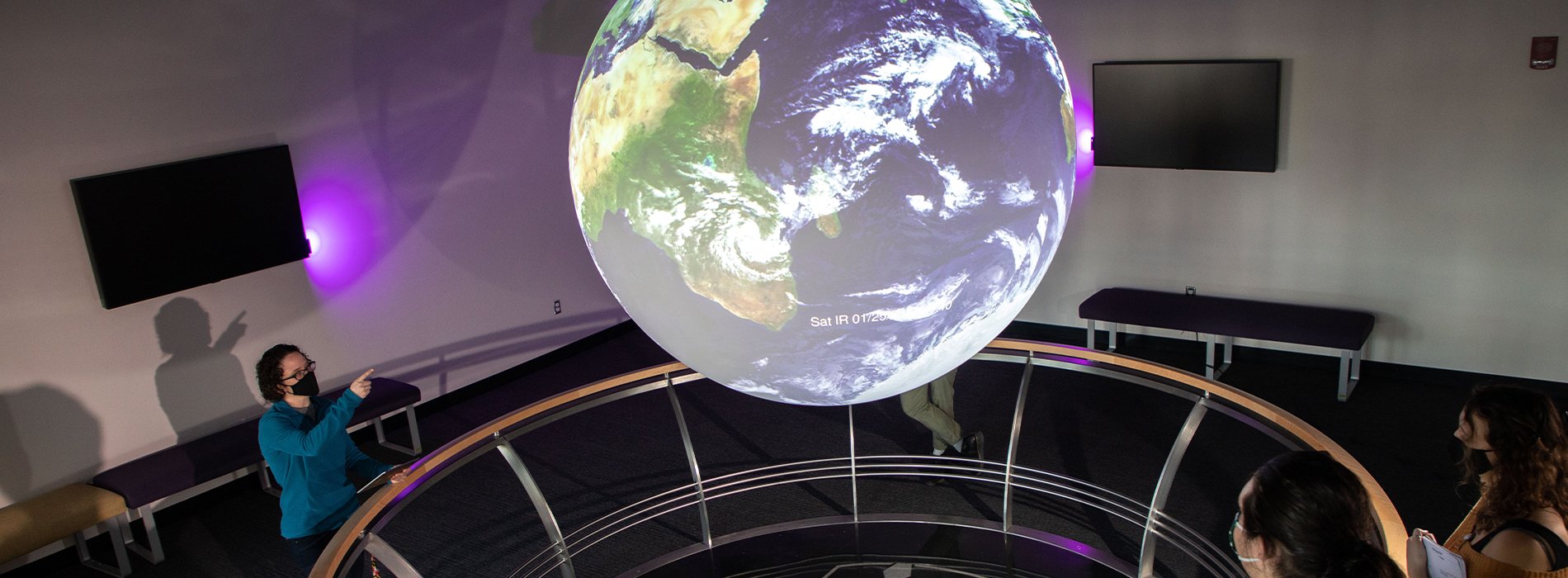 Four people observe a sphere displaying a satellite projection of the earth in UAlbany's Science-On-A-Sphere room