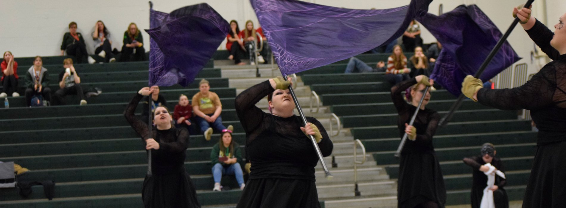 The UAlbany Winter Guard performs with all black costumes and purple flags inside a gymnasium.