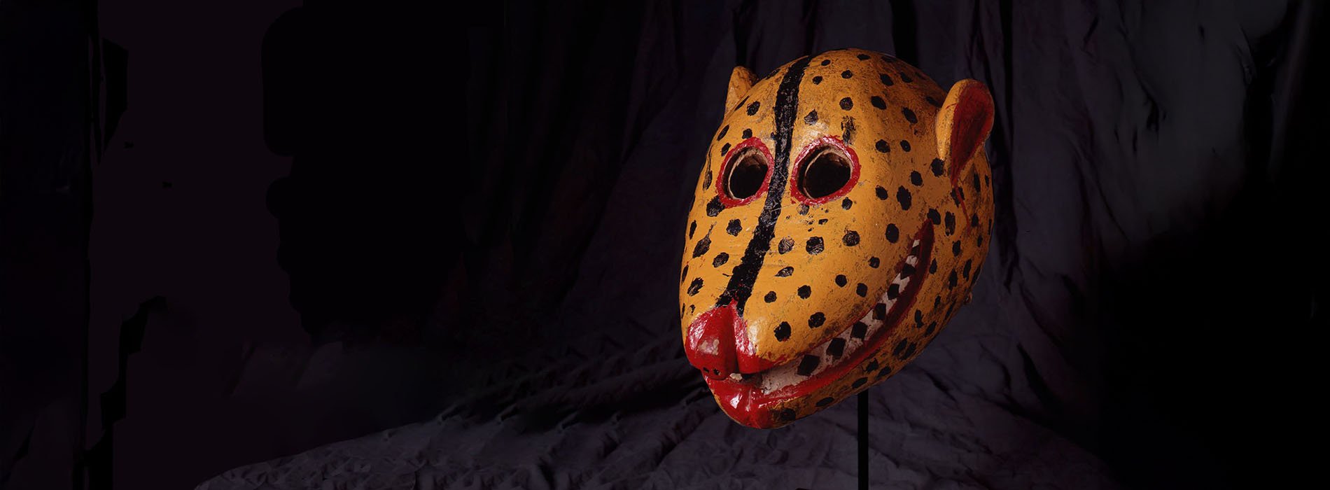 Yellow, red and black jaguar mask on a stand with a dark cloth backdrop