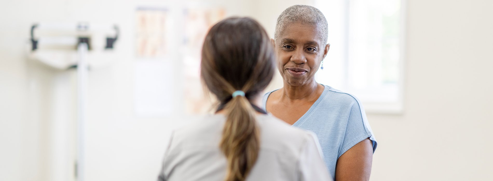 A black woman in a loose blue shirt and cropped gray hair smiles at a doctor with brown hair in a ponytail.