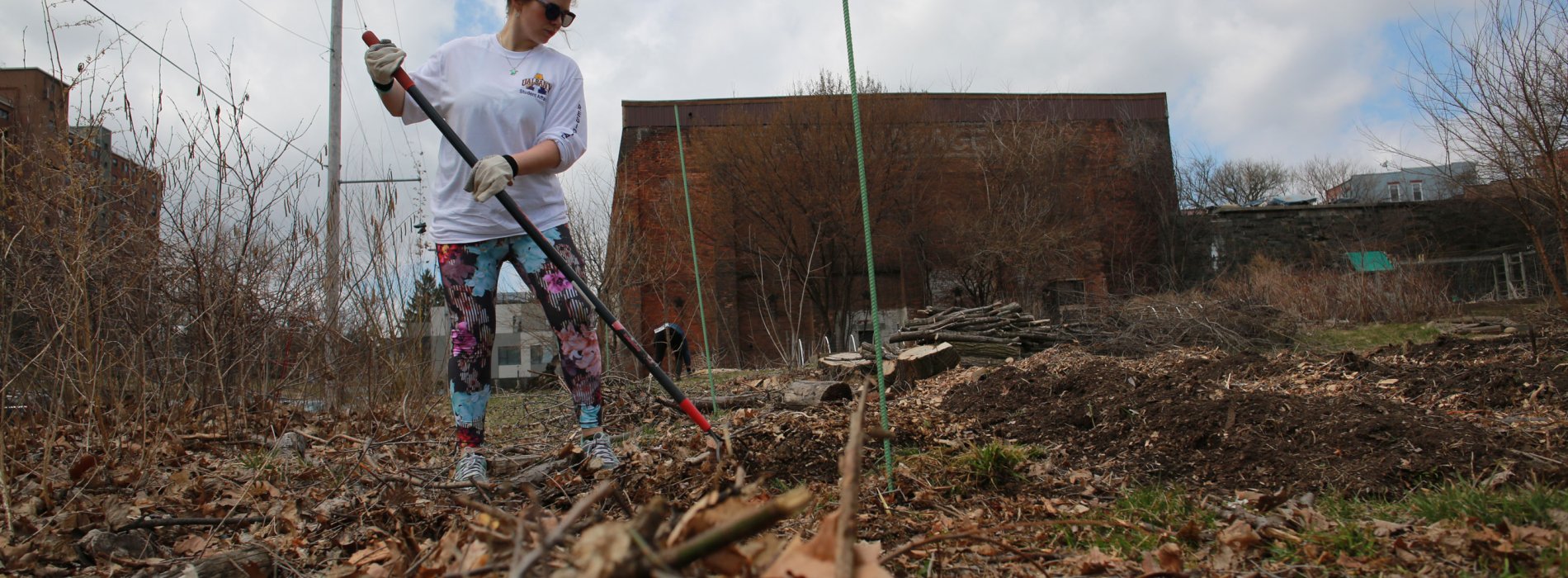 A student rakes debris in an empty urban lot on a cloudy day.