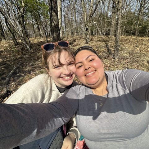 Two young women stand together, smiling for a selfie, in a forest in early spring. 