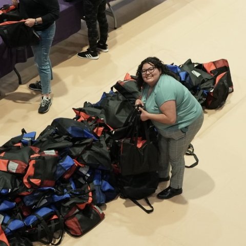 A woman with dark curly hair and glasses stands beside a large pile of blue and red duffle bags.