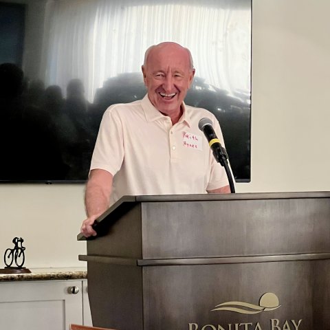 A man in a white short sleeved polo shirt smiles at a dark wooden lectern. 