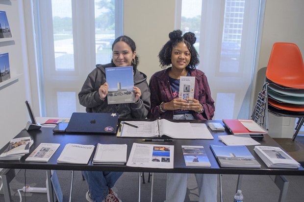 Two students sitting at a desk holding up brochures and smiling. The desk has additional brochures on it.